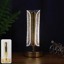 A lit gold and clear crystal table lamp on a dark wooden surface, with a small cannon and plant in the background.