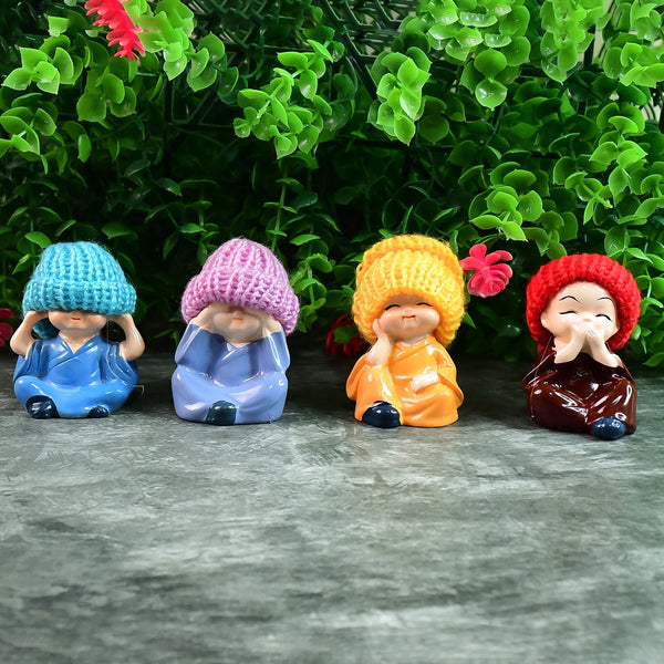 Four resin baby monk figurines with colorful knitted hats sitting on a gray surface in front of green foliage. The first