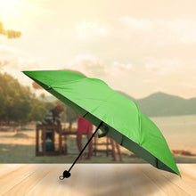 An open green and silver umbrella with a black handle rests on a wooden surface with a beach background.