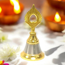 A small brass and steel pooja bell with a peacock feather design stands on a white surface surrounded by lit diyas and white