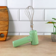 Light green cordless electric whisk standing upright on a brown counter, next to a wooden cutting board and a plant.