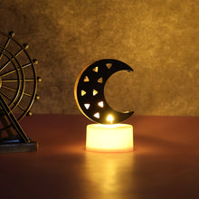 A single crescent moon LED tea light with warm yellow glow on a dark table next to a decorative metal Ferris wheel.