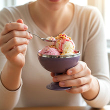 A person holds a purple dessert bowl containing two scoops of ice cream, taking a spoonful.