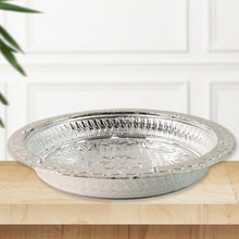 A silver-colored plastic pooja thali with embossed patterns, sitting on a light wooden surface with a white paneled wall