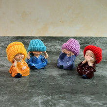 Four resin baby monk figurines with colorful knitted hats sitting on a gray surface against a light gray wall. The first