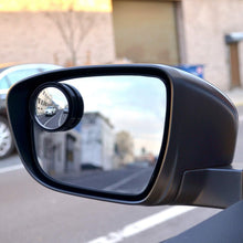 A car's side mirror with a small, round blind spot mirror attached, reflecting a city street.