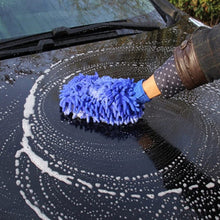 A hand wearing a blue microfiber wash mitt creates circular soap patterns on a black car hood.