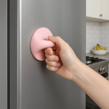 A hand pulls a pink self-adhesive handle attached to a gray refrigerator in a kitchen.