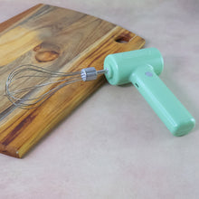Light green cordless electric whisk with its attachment resting on a brown counter next to a wooden cutting board.