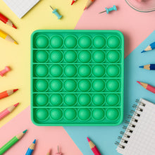 A square green silicone pop-it fidget toy surrounded by colorful pencils and thumbtacks on a multi-colored background.