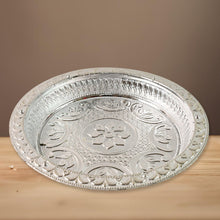 A silver-colored plastic pooja thali with embossed floral designs, resting on a light wooden surface with a brown background.