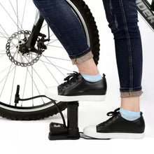 A person in blue jeans and black sneakers uses a black foot pump to inflate a bicycle tire.