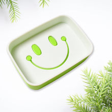 A close-up of a green and white soap dish with a prominent green smiley face draining design.