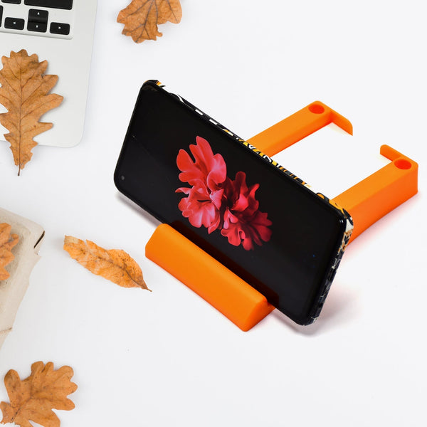 An orange plastic holder on a white desk, holding a black smartphone horizontally, surrounded by autumn leaves.