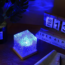 A crystal cube lamp projecting blue ripples on a desk with a calendar, books, and a plant in the background.