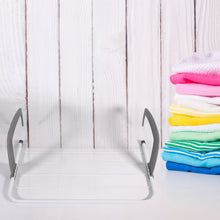 White and gray clothes drying rack on a white surface with a stack of colorful folded clothes to the right.