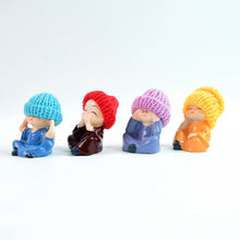 Four resin baby monk figurines with colorful knitted hats sitting on a white surface. The first monk wears a blue hat and