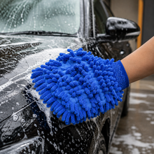 A hand wearing a blue microfiber wash mitt cleans a black car with white soap suds.