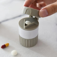 A hand opening a gray and white pill cutter with a white pill inside and a capsule nearby.