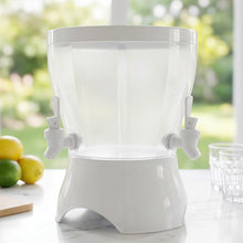 An empty, clear plastic dual-compartment beverage dispenser with a white base and two taps sits on a marble countertop with