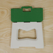 A green and white plastic folding stool is shown partially folded, revealing its carry handle, on a wooden surface.