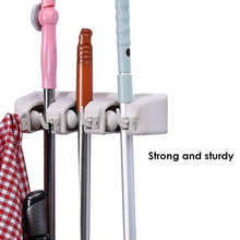 Close-up of the white three-slot wall-mounted holder, holding a pink broom, a brown broom, and a blue mop, with a red
