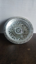 A silver-colored plastic pooja thali with embossed patterns, on a dark brown surface with a white textured wall in the