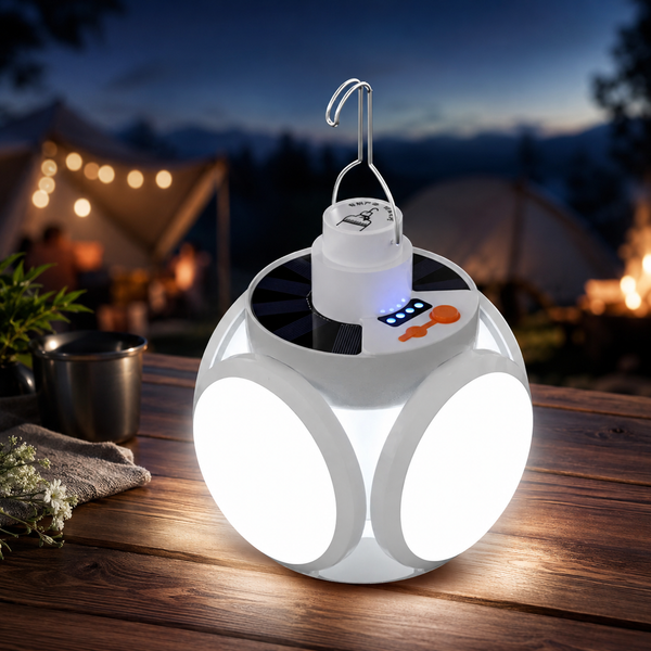 A white solar camping light with four illuminated panels sits on a wooden table at dusk, with tents in the background.