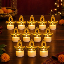 Twelve golden diya LED tealight candles, with white bases, glowing on a dark wooden surface with flowers in the background.