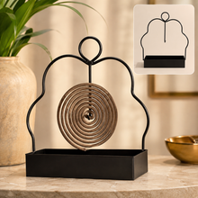 A black metal mosquito coil holder with a brown spiral coil sits on a marble surface next to a plant.