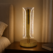 A lit gold and clear crystal table lamp with a curved design sits on a nightstand next to a bed.