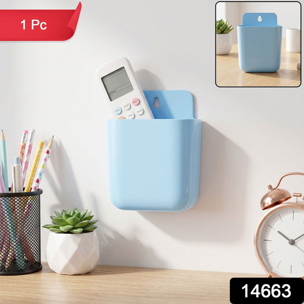 A light blue wall-mounted plastic storage pocket holds a white remote control above a desk with a plant and clock, with a "1