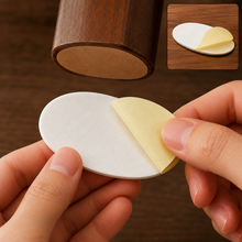 Hands peeling back the yellow adhesive layer from a white oval pad, with a furniture leg above.