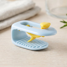 A single blue and yellow plastic clip rests on a light wooden surface, with a blurred background of kitchen items.