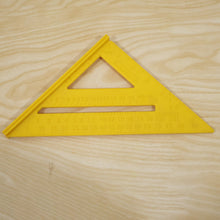 A yellow plastic speed square ruler with black markings is placed on a light wood grain surface.