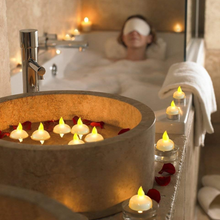 A stone basin filled with water, red petals, and white LED floating candles, with a woman bathing in the background.