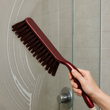 A hand uses a red and white handled brush with brown bristles to scrub a soapy shower glass door.