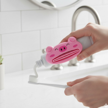 Hands using a pink pig-shaped toothpaste squeezer to dispense white toothpaste onto a white toothbrush over a bathroom sink.
