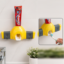 Yellow and gray toothpaste dispenser mounted on a white tiled wall with a tube of toothpaste inserted, and a hand holding a