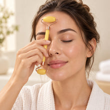 A woman with closed eyes gently rolls a yellow jade facial massager across her forehead in a bright bathroom setting.