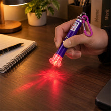 A hand holds a purple laser pointer keychain, projecting a red light onto a wooden desk beside a notebook.