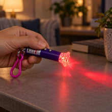 A hand holds a purple laser pointer keychain, casting a red light pattern onto a grey tabletop in a dimly lit room.