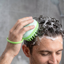 A person's hand is visible, holding a light green silicone scalp massage brush and scrubbing their dark, sudsy hair in a