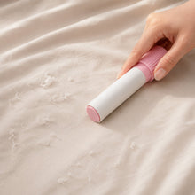 A hand uses a pink and white lint roller on a light beige fabric surface, removing lint and fuzz.