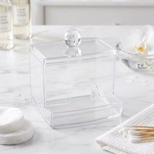 Empty clear acrylic cotton swab dispenser on a white marble counter with other toiletries.