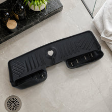 A black silicone faucet splash guard with drainage grooves and two side compartments, lying on a light grey tiled floor next