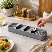 A hand places a silver fork into a gray plastic cutlery organizer on a white marble counter with food and plants.