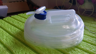 A translucent collapsible water container with a blue spigot and white handle rests on a textured green surface indoors.