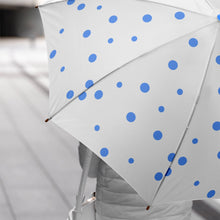 A person holds an open white umbrella with blue polka dots, walking on a grey sidewalk.