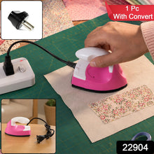 A hand holds a pink and white mini iron on fabric, with a black converter plug and a red banner saying 